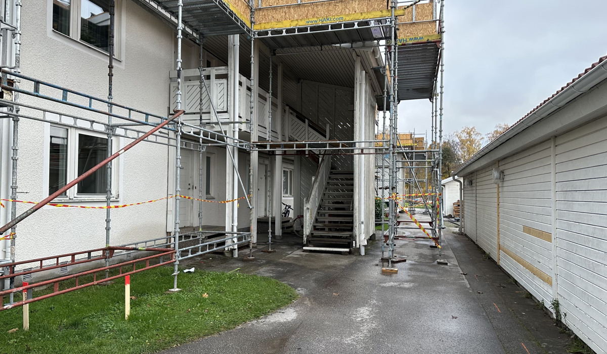 Kortsidan av ett tvåvåningshus i vitt trä är täckt av byggställningar inför en renovering. Mittemot byggnaden syns en sidobyggnad där man byter ut delar av fasaden. 