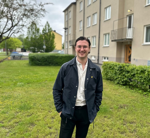 En glad man med mörka kläder framför ett ljust trevåningshus
