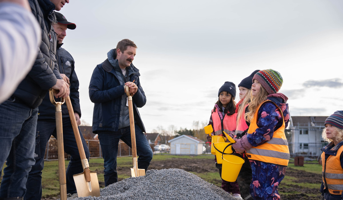 Tre män i 40-50-årsåldern står på en grushög med varsin förgylld spade i händerna. Mittemot dem står fyra femåringar med gula plasthinkar och spade.